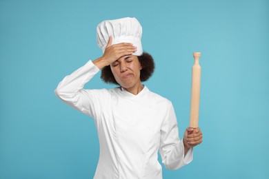 Photo of Frustrated female chef in uniform holding rolling pin on light blue background