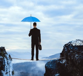 Image of Risks and challenges of entrepreneurship. Businessman with portfolio and umbrella standing on rope over abyss in mountains, back view