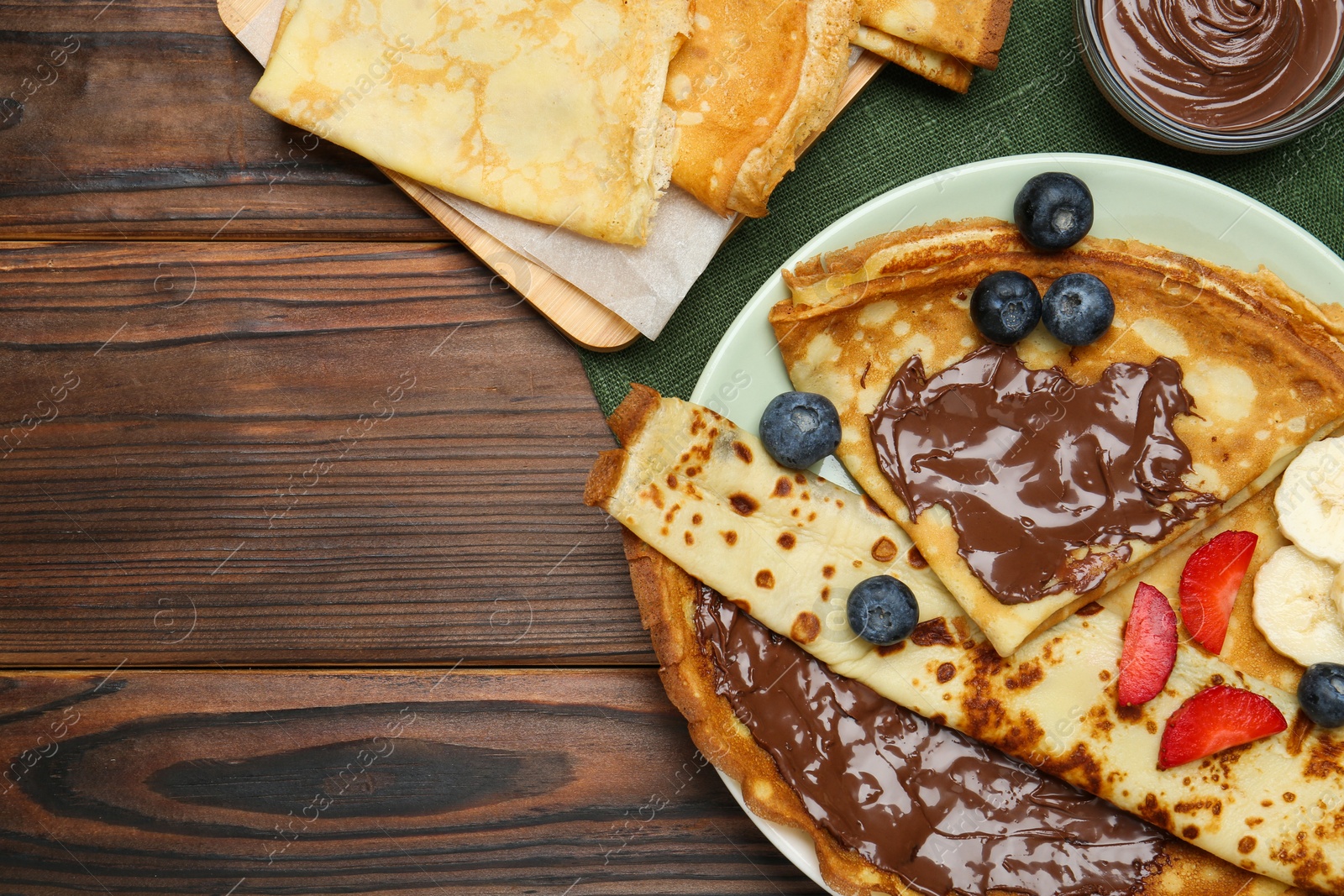 Photo of Tasty crepes with chocolate paste, banana and berries served on wooden table, flat lay. Space for text