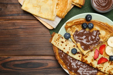 Tasty crepes with chocolate paste, banana and berries served on wooden table, flat lay. Space for text