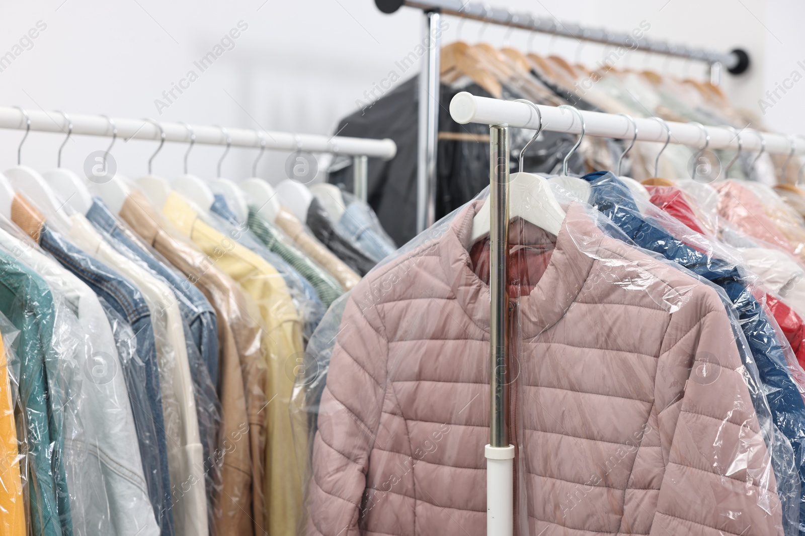 Photo of Dry-cleaning service. Many different clothes in plastic bags hanging on rack indoors, closeup