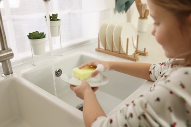 Photo of Little girl washing plate above sink indoors, closeup