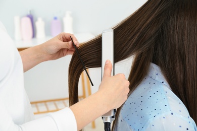 Photo of Hairdresser using modern flat iron to style client's hair in salon, closeup