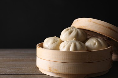 Photo of Delicious bao buns (baozi) in bamboo steamer on wooden table against black background, closeup. Space for text