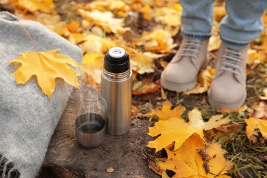 Photo of Woman standing near metallic thermos, cap and scarf on tree stump in autumn park, closeup