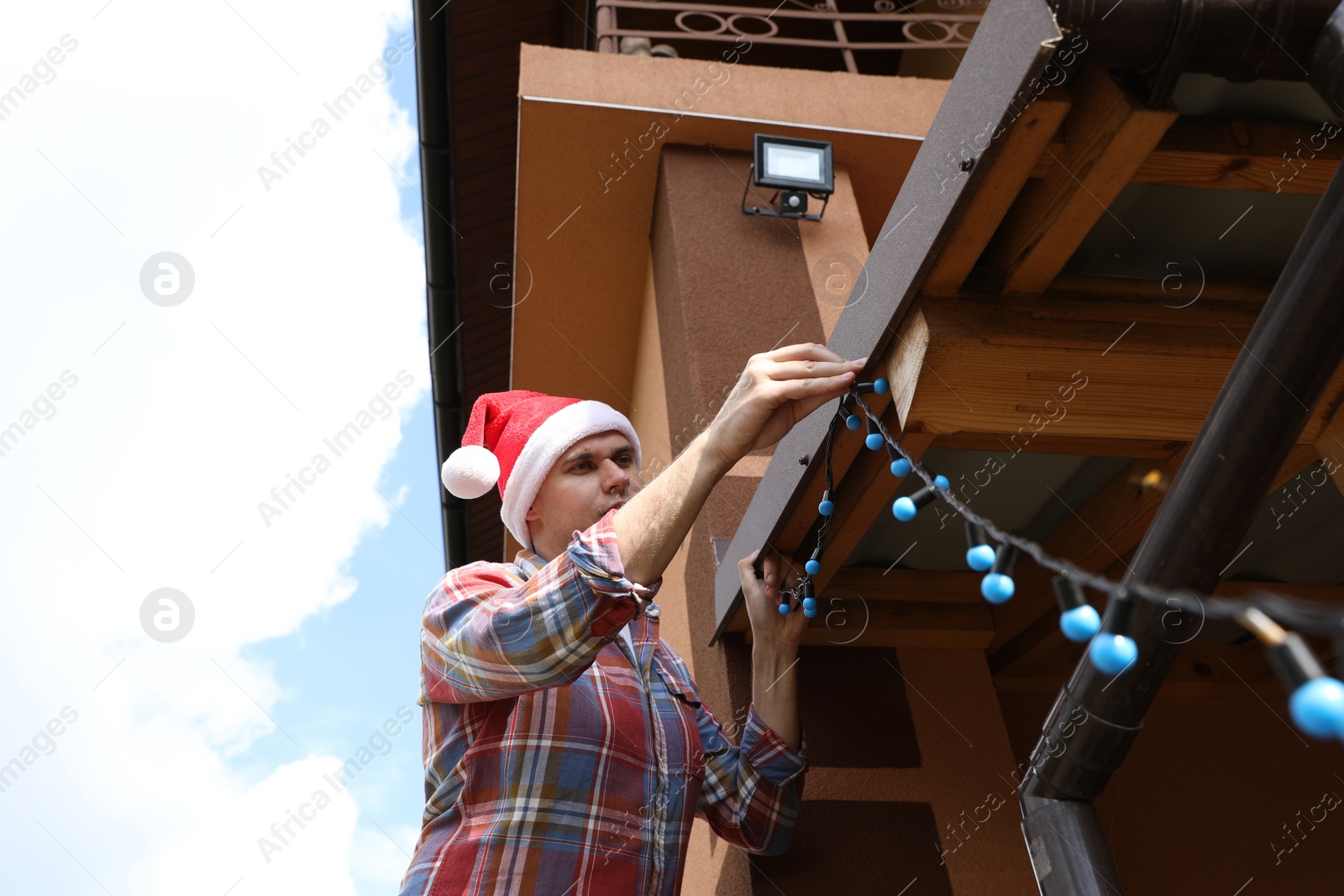 Photo of Man in Santa hat decorating house with Christmas lights outdoors, low angle view