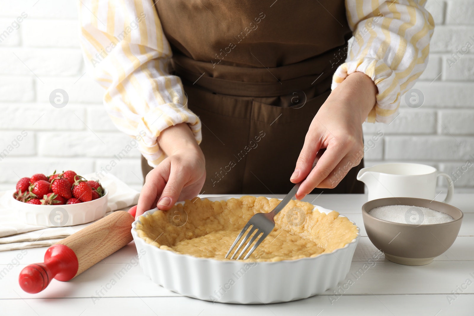 Photo of Shortcrust pastry. Woman making holes in raw dough with fork at white wooden table, closeup