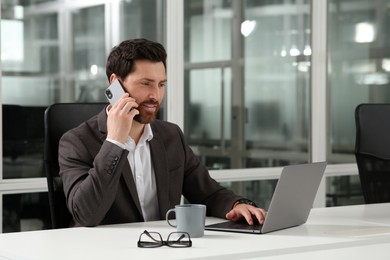 Man talking on phone while working with laptop at white desk in office
