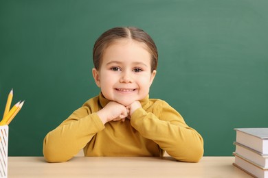 Happy little school child sitting at desk with books near chalkboard