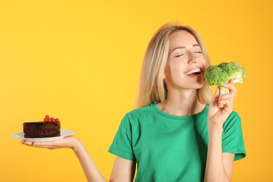 Woman choosing between cake and healthy broccoli on yellow background