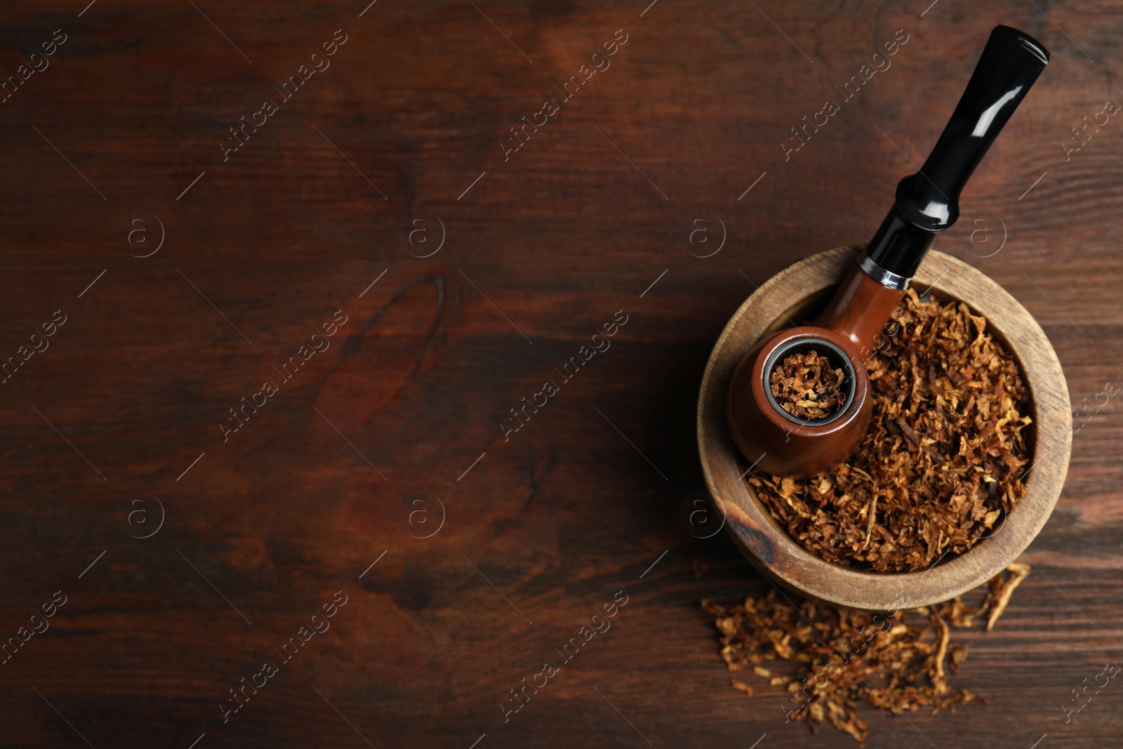 Photo of Smoking pipe and bowl of dry tobacco on wooden table, top view. Space for text