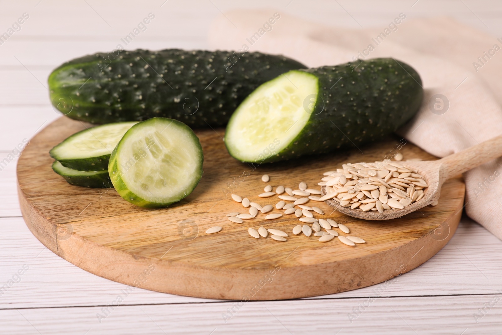 Photo of Spoon of vegetable seeds and fresh cucumbers on white wooden table, closeup