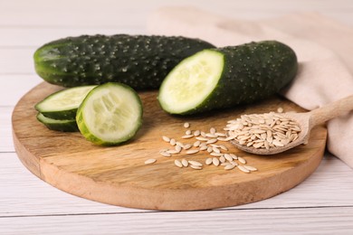 Photo of Spoon of vegetable seeds and fresh cucumbers on white wooden table, closeup