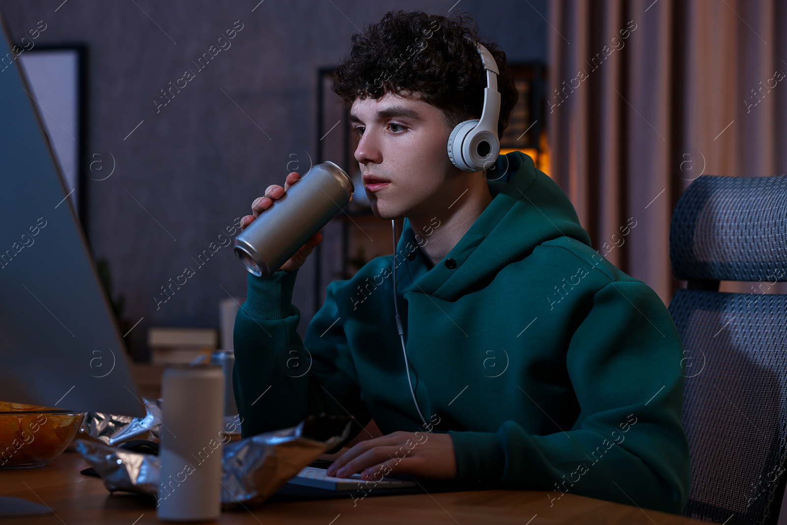 Photo of Young man with energy drink and headphones playing video game at wooden desk indoors