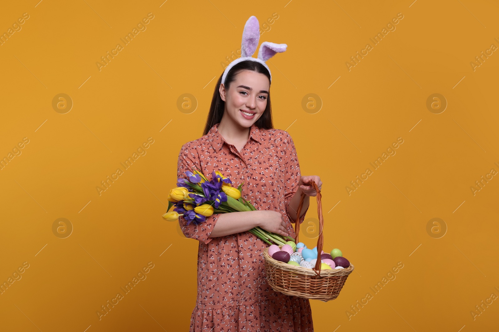 Photo of Happy woman in bunny ears headband holding wicker basket with painted Easter eggs and bouquet of flowers on orange background