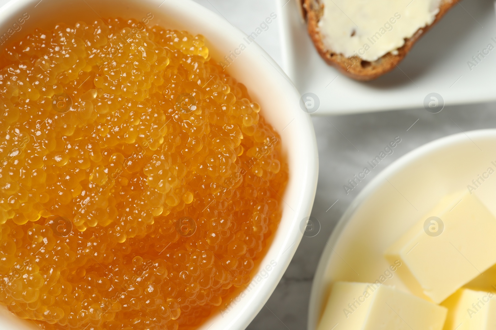 Photo of Fresh pike caviar in bowl, bread and butter on marble table, top view