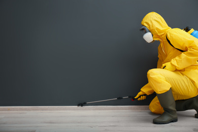 Photo of Pest control worker in protective suit spraying pesticide near black wall indoors. Space for text