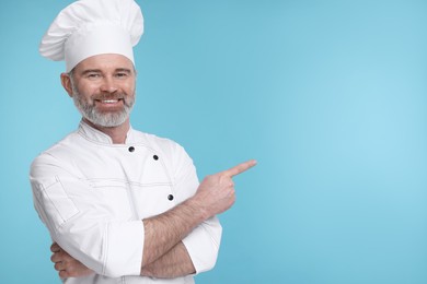 Photo of Happy chef in uniform pointing at something on light blue background, space for text