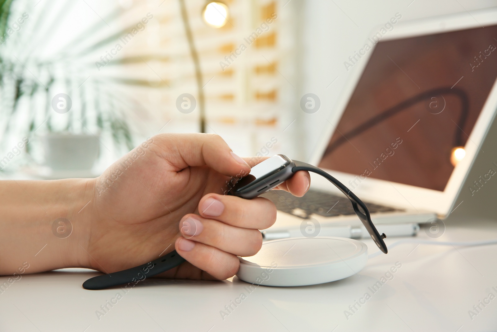 Photo of Man taking smartwatch from wireless charger at white table, closeup. Modern workplace accessory