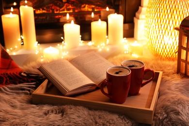 Photo of Cups of hot drink and open book near fireplace at home