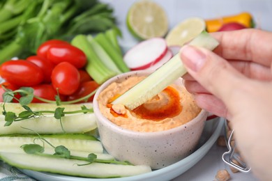 Photo of Woman dipping cucumber stick into hummus at table, closeup