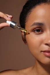 Woman applying serum onto her face on brown background, closeup