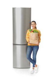 Photo of Young woman with bag of groceries near closed refrigerator on white background