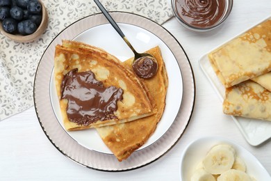 Tasty crepes with chocolate paste, blueberries and banana served on white wooden table, flat lay