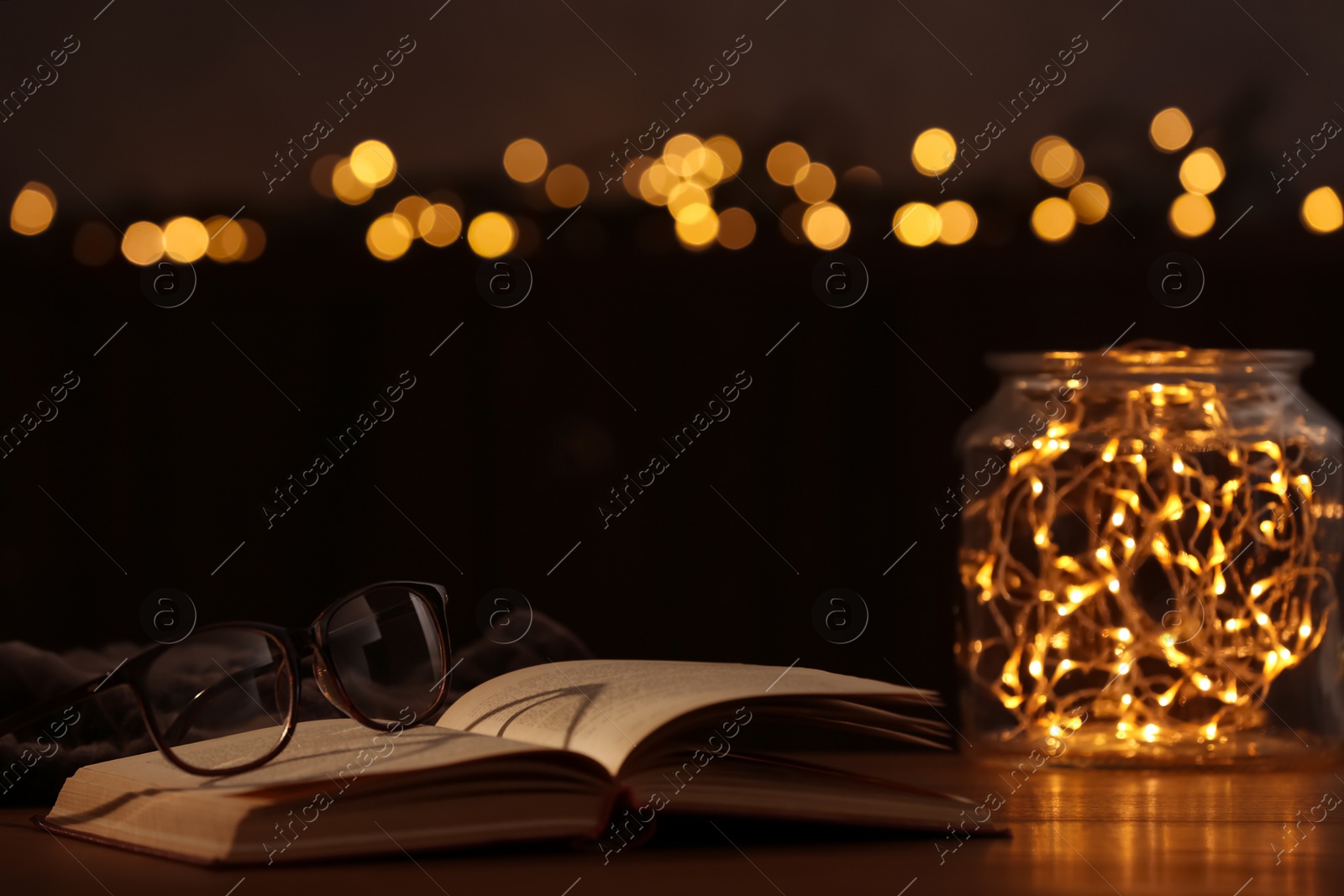 Photo of Eyeglasses and open book on wooden table against festive lights. Space for text