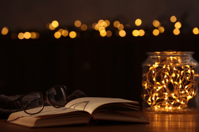 Photo of Eyeglasses and open book on wooden table against festive lights. Space for text