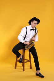 Young man in elegant outfit playing saxophone on yellow background