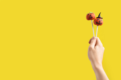 Photo of Woman with delicious pumpkin shaped cake pops and space for text on yellow background, closeup. Halloween celebration