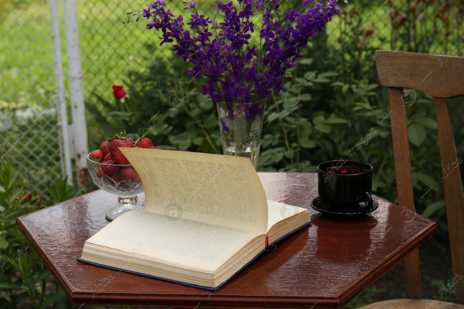 Photo of Open book, cup of tea, strawberries and beautiful wildflowers on table in garden