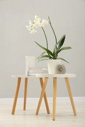 Photo of Blooming white orchid flower in pot, books and candle on nesting tables near grey wall indoors