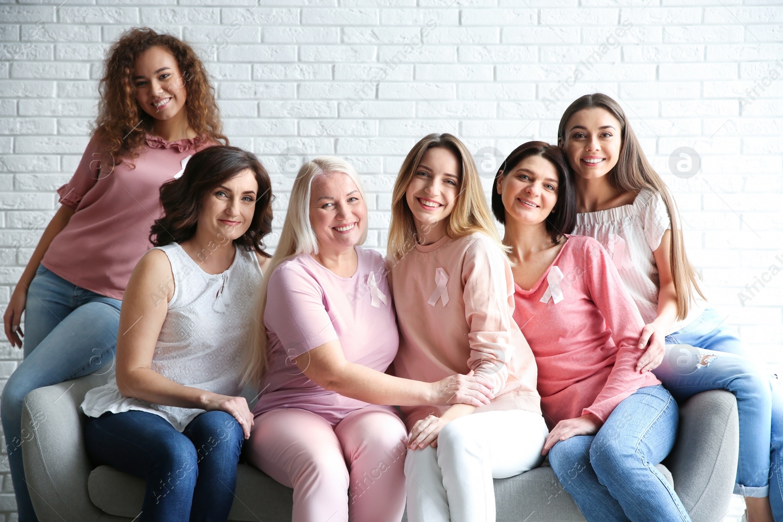 Photo of Group of women with silk ribbons sitting on sofa against brick wall. Breast cancer awareness concept
