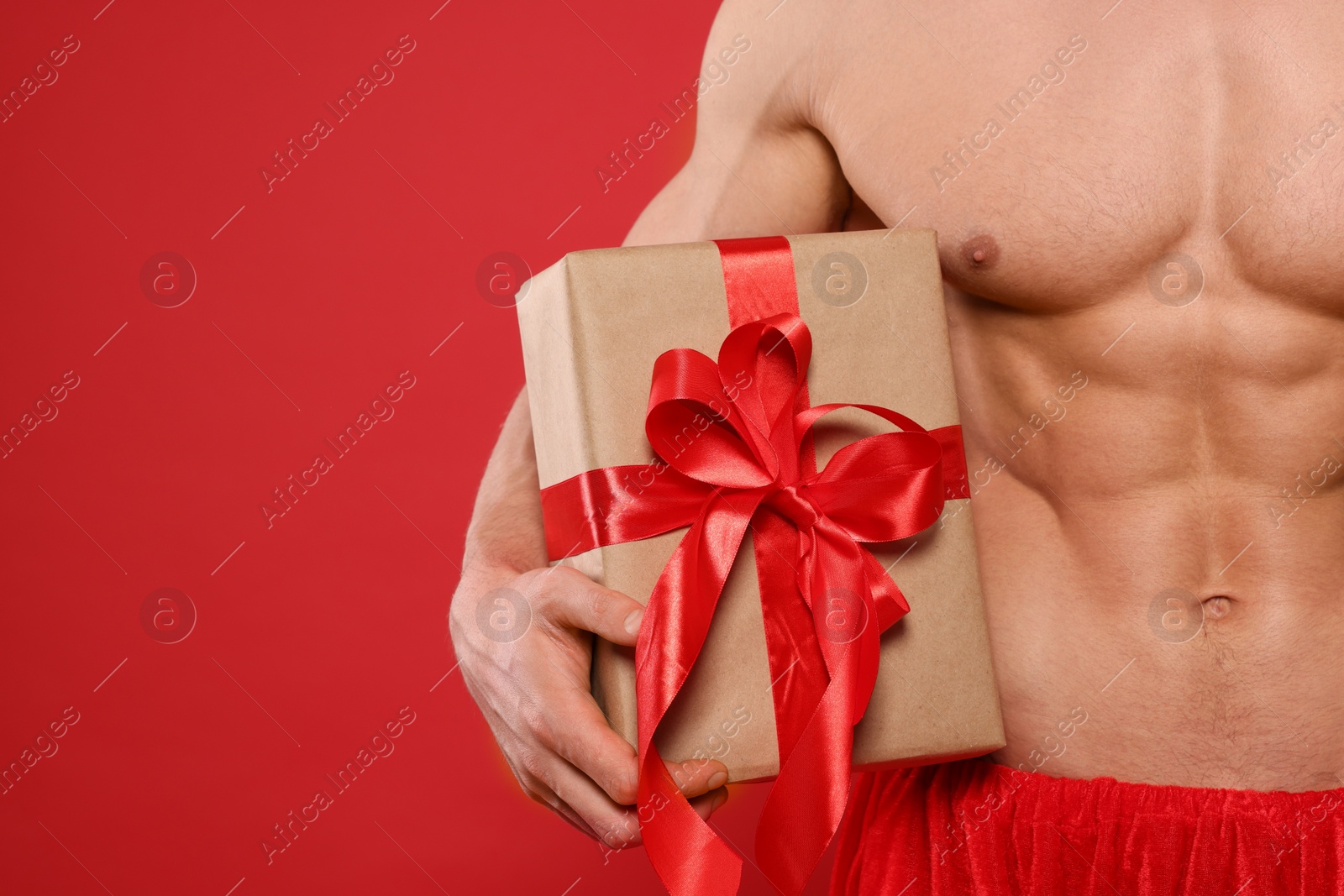 Photo of Young man with muscular body holding Christmas gift box on red background, closeup. Space for text