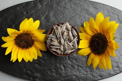 Raw sunflower seeds and flowers on slate board, flat lay