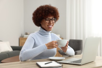 Photo of Beautiful young woman using laptop and drinking coffee at wooden desk in room