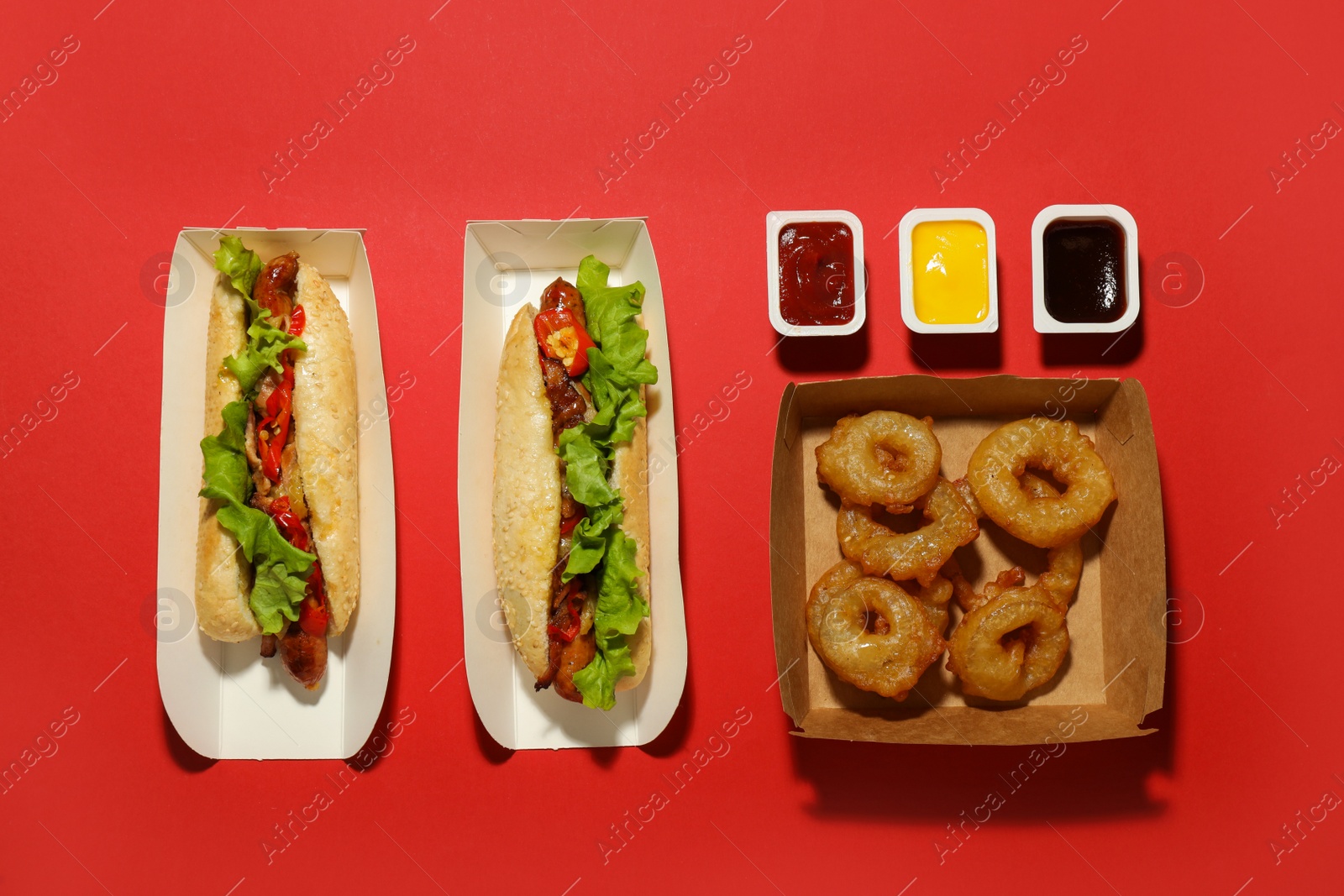 Photo of Tasty hot dogs, fried onion rings and sauces on red background, flat lay. Fast food