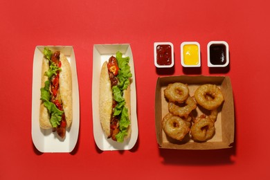 Photo of Tasty hot dogs, fried onion rings and sauces on red background, flat lay. Fast food