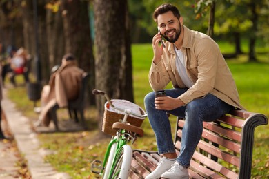 Young man sitting on bench with coffee and talking over smartphone in park, space for text