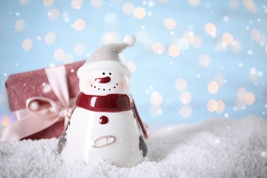 Decorative snowman near gift box on artificial snow against blurred festive lights, space for text