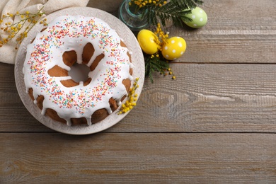Photo of Glazed Easter cake with sprinkles, painted eggs and flowers on wooden table, flat lay. Space for text