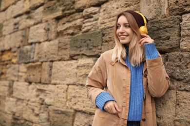 Young woman with headphones listening to music near stone wall. Space for text
