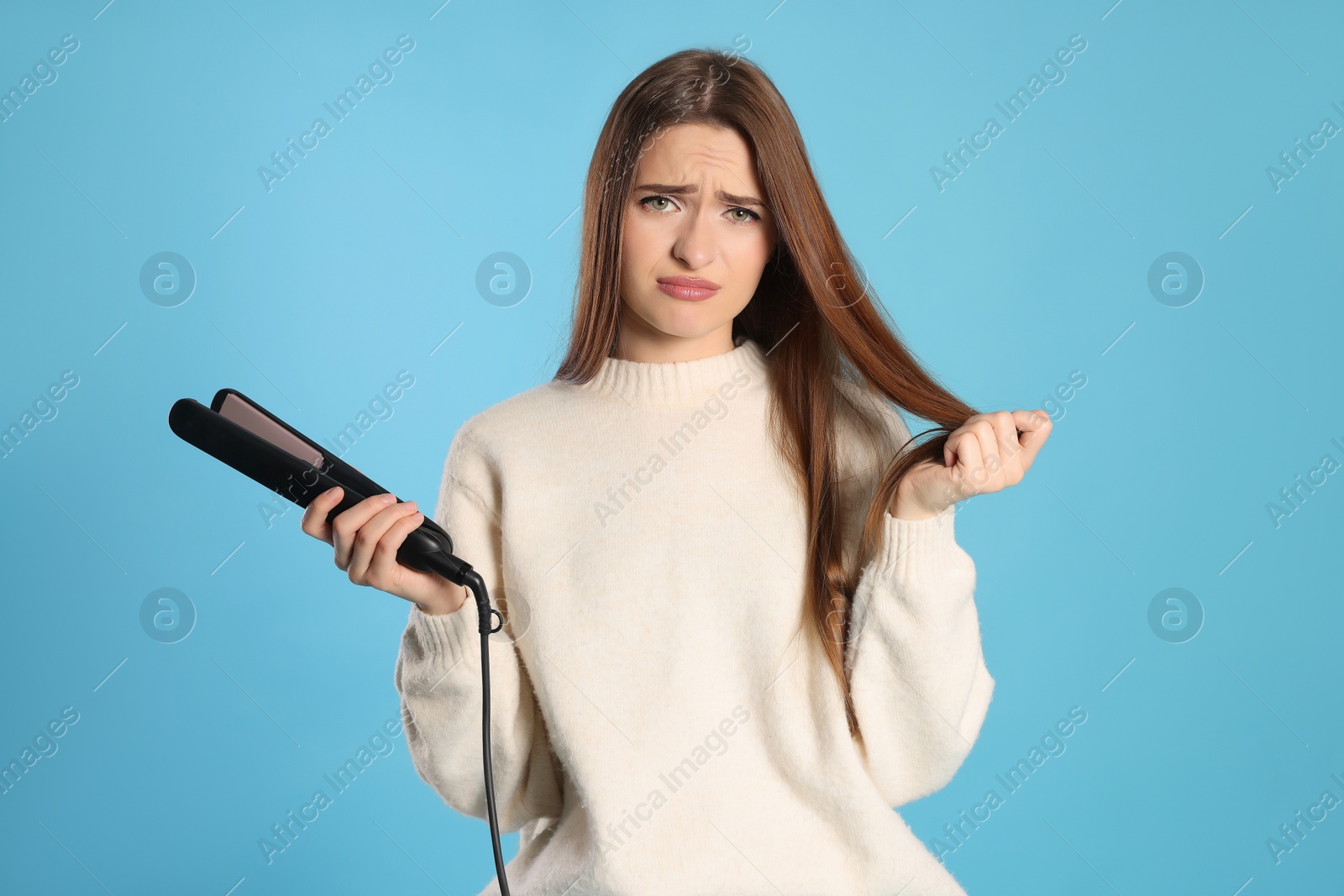 Photo of Upset young woman with flattening iron on light blue background. Hair damage