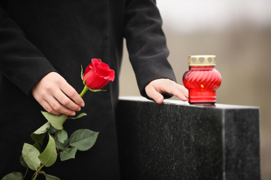 Photo of Woman holding red rose near black granite tombstone with candle outdoors, closeup. Funeral ceremony