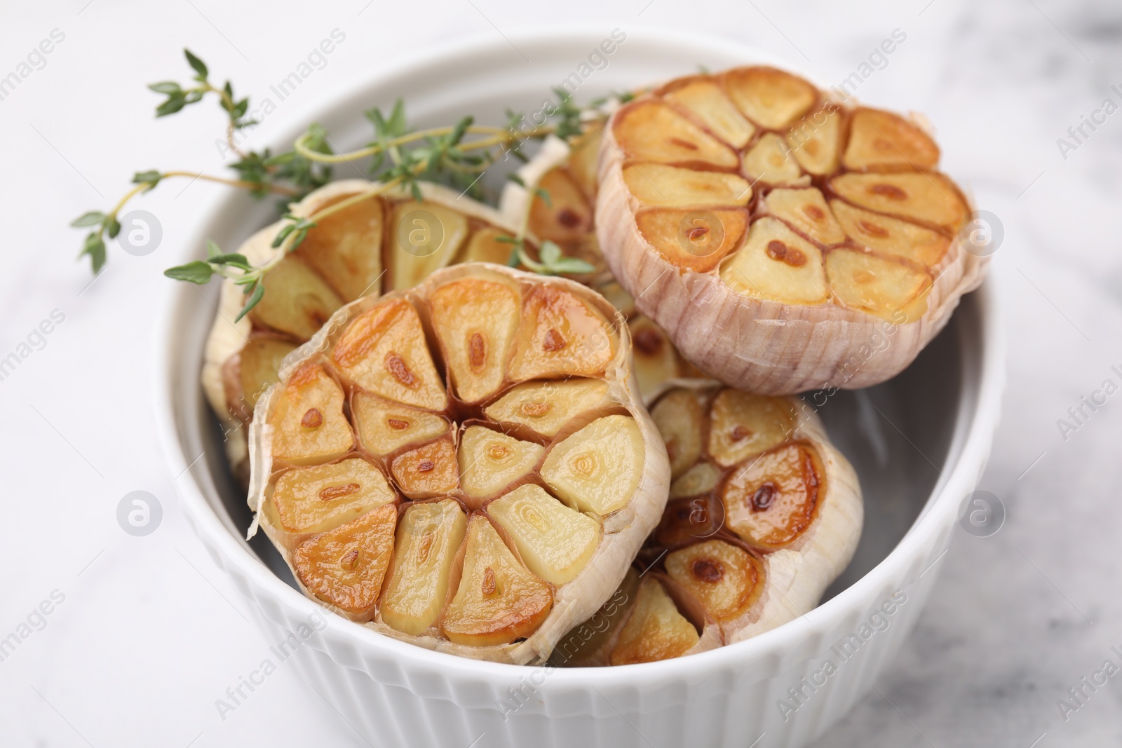 Photo of Heads of fried garlic and thyme in bowl on white table, closeup