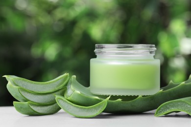 Photo of Jar with cream and cut aloe leaf on white table against blurred green background, closeup