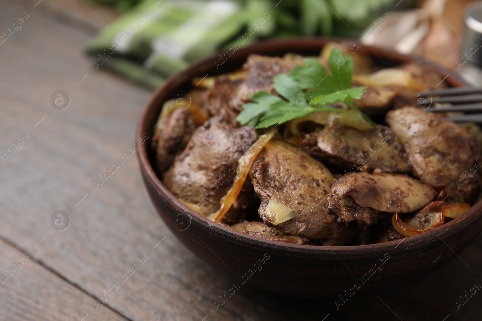 Photo of Tasty fried chicken liver with onion and parsley in bowl on wooden table, closeup