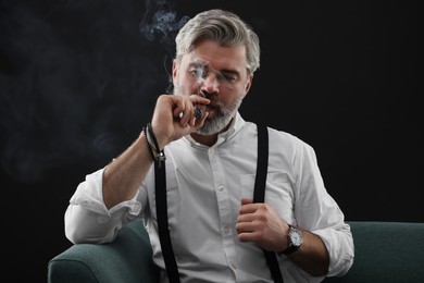 Bearded man smoking cigar on sofa against black background